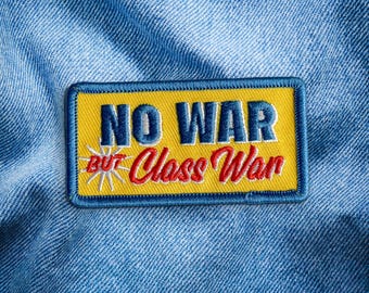 May include: Embroidered patch with the text "NO WAR BUT Class War" in blue and red on a yellow background. The patch has a blue border and a starburst graphic. The patch is on a blue denim background.