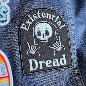 May include: Black and white embroidered patch with a skeleton wearing a hooded robe and making the 'rock on' hand gesture. The patch says "Existential Dread" in white text.
