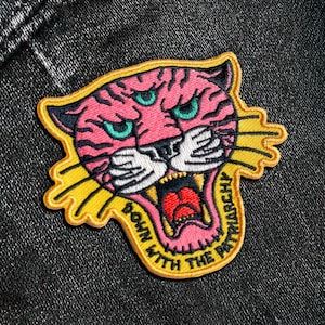 May include: Embroidered patch featuring a pink tiger head with three eyes, black stripes, and teal eyes. The tiger's mouth is open, revealing a red tongue and teeth. The patch has the text "DOWN WITH THE PATRIARCHY" and a yellow border.