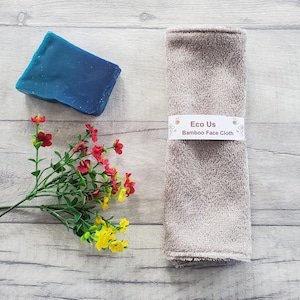May include: A light brown bamboo face cloth with the text "Eco Us Bamboo Face Cloth" on a white label. The cloth is on a light wood surface with a blue bar of soap and a small bouquet of red and yellow flowers.