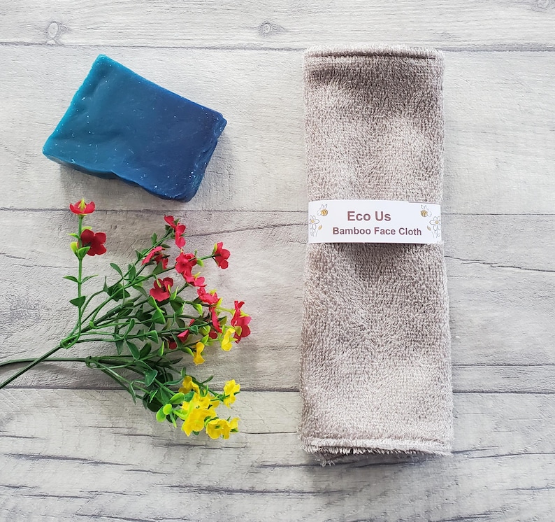 May include: A light brown bamboo face cloth with the text "Eco Us Bamboo Face Cloth" on a white label. The cloth is on a light wood surface with a blue bar of soap and a small bouquet of red and yellow flowers.