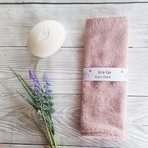 May include: A pink face cloth with the text "Eco Us Face Cloth" printed on a white label. The cloth is folded and sitting on a light wood surface. There is a white bar of soap and a sprig of lavender flowers in the image.