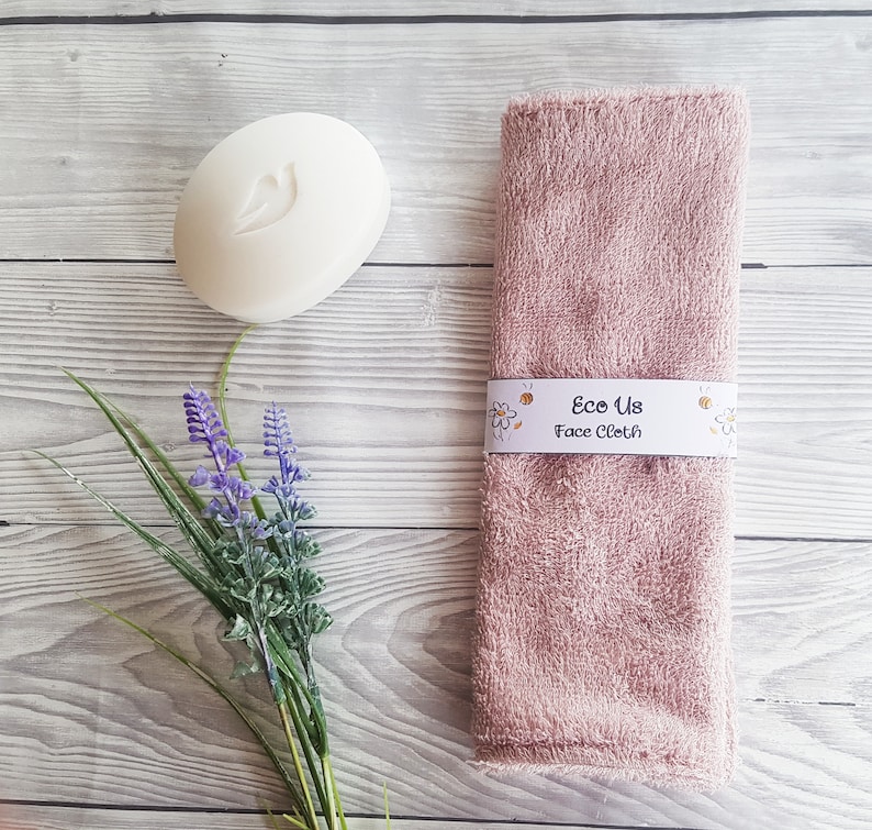 May include: A pink face cloth with the text "Eco Us Face Cloth" printed on a white label. The cloth is folded and sitting on a light wood surface. There is a white bar of soap and a sprig of lavender flowers in the image.