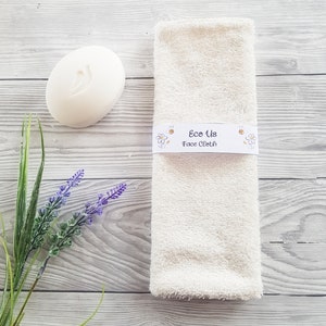 May include: A white face cloth with the text "Eco Us Face Cloth" printed on a label. The label has a bee and daisy design. The cloth is on a light wood background with a white bar of soap and lavender flowers.