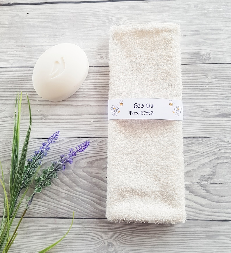 May include: A white face cloth with the text "Eco Us Face Cloth" printed on a label. The label has a bee and daisy design. The cloth is on a light wood background with a white bar of soap and lavender flowers.