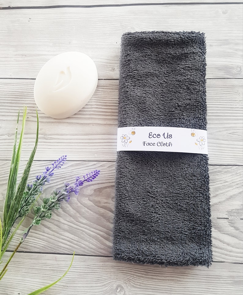 May include: A gray face cloth with the text "Eco Us Face Cloth" on a white label. The cloth is on a wooden surface with a white bar of soap and a sprig of lavender.