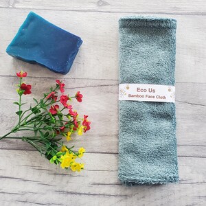 May include: A light blue bamboo face cloth with the text "Eco Us Bamboo Face Cloth" on a white label. The cloth is on a wooden surface with a blue bar of soap and a bouquet of red and yellow flowers.