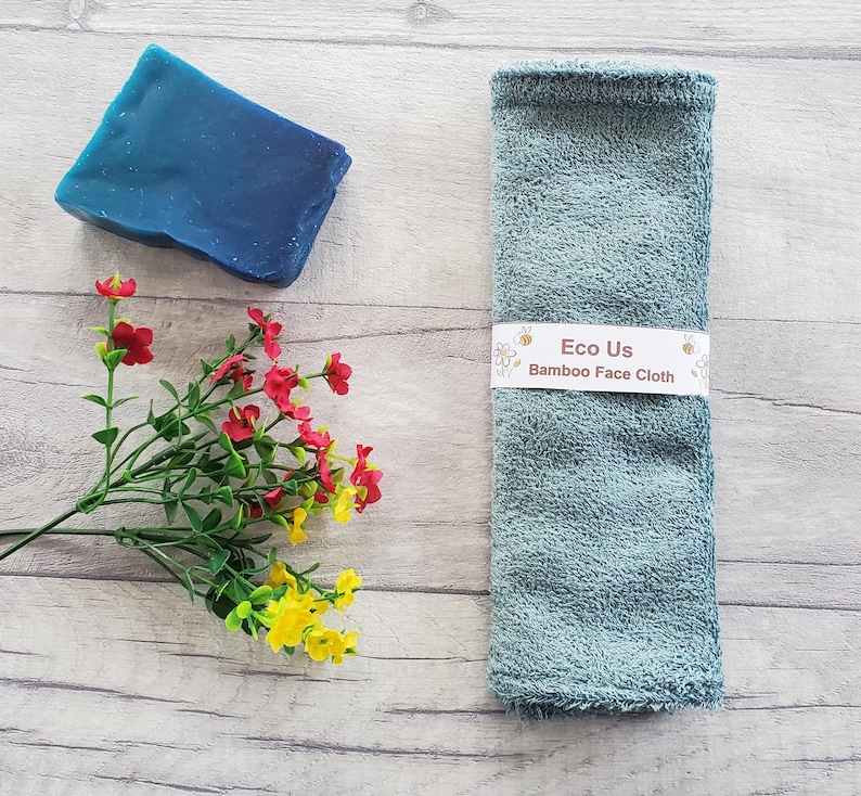 May include: A light blue bamboo face cloth with the text "Eco Us Bamboo Face Cloth" on a white label. The cloth is on a wooden surface with a blue bar of soap and a bouquet of red and yellow flowers.