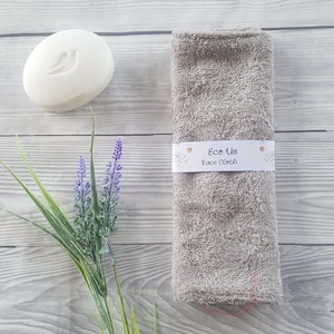 May include: A gray face cloth with the text "Eco Us Face Cloth" on a white label. The cloth is folded and sitting on a wooden surface with a white bar of soap and lavender flowers.