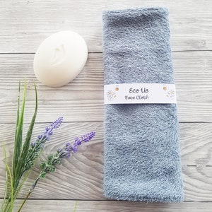 May include: A light blue face cloth with the text "Eco Us Face Cloth" on a white label. The cloth is folded and sitting on a wooden surface with a white bar of soap and a sprig of lavender.