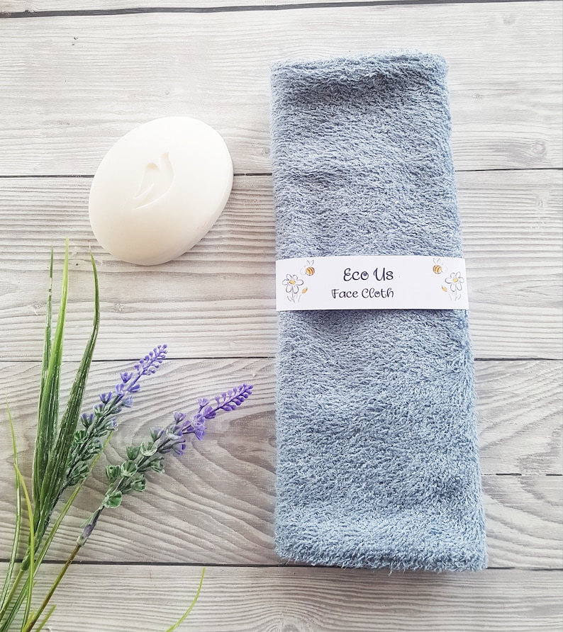 May include: A light blue face cloth with the text "Eco Us Face Cloth" on a white label. The cloth is folded and sitting on a wooden surface with a white bar of soap and a sprig of lavender.
