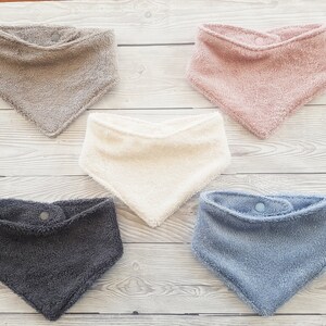 Pode incluir: Cinco babadores de tecido triangulares com fechos de pressão. Os babadores são bege, rosa, creme, cinzento e azul.
