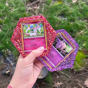 Puede incluir: Dos posavasos hexagonales de resina, uno rosa y otro morado, cada uno con una carta coleccionable de Pokémon. El posavasos rosa presenta una carta de Sylveon, mientras que el morado muestra una carta de Espeon. Ambos posavasos están cubiertos de purpurina.