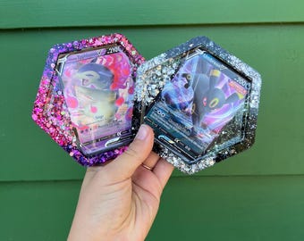 May include: Two hexagonal resin Pokemon card holders with glitter embedded in the resin. The card holders are black and white with silver glitter and pink and purple with silver glitter. The cards inside the holders are for the Pokemon Typhlosion V and Umbreon V.