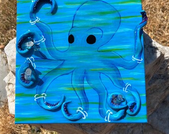 May include: A blue octopus with white eyes and purple accents is painted on a blue and green striped canvas. The octopus has eight tentacles with crystals embedded in each tentacle.
