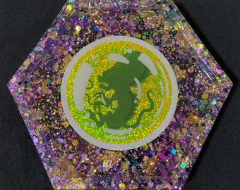 May include: Hexagon-shaped resin coaster with a green creature in a bubble design. The coaster is filled with iridescent glitter and gold flakes.