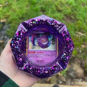 Puede incluir: Cenicero de resina con purpurina morada con una carta de Pokémon Gengar en su interior. La carta muestra la habilidad de Gengar, Life Shaker, y su ataque, Hypnoblast.