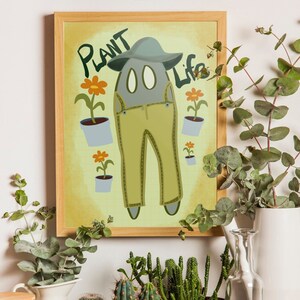 Puede incluir: Una impresión enmarcada con un fondo amarillo y un personaje de dibujos animados que lleva un mono y un sombrero. El personaje está rodeado de flores y el texto "Plant Life" está en la parte superior de la impresión.