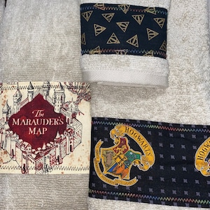 Peut inclure: Trois serviettes blanches avec des bordures en tissu noir et or. La serviette du haut a une bordure en tissu noir avec un motif de triangles dorés. La serviette du milieu a une bordure en tissu avec une illustration rouge et blanche du château de Poudlard et le texte "The Marauder's Map". La serviette du bas a une bordure en tissu noir avec un motif de diamants noirs et l'emblème de Poudlard avec le texte "Hogwarts".