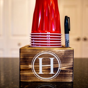 May include: A wooden cup holder with a white letter "H" in a circle on the front. The holder has a slot for a marker and is filled with red plastic cups.
