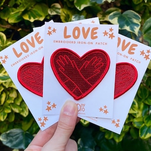 May include: Three red embroidered iron-on patches in the shape of a heart with two hands inside. The patches are on white cards with the text "LOVE EMBROIDERD IRON-ON PATCH" and stars.