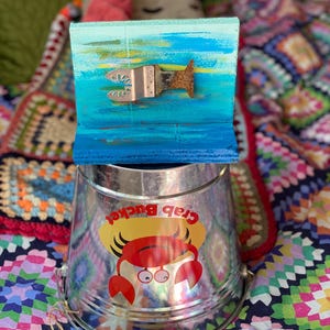 May include: A silver metal crab bucket with a red crab illustration and the text "Crab Bucket". The bucket is sitting on a colourful blanket with a blue and green painted wooden sign behind it.