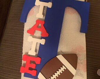 Könnte beinhalten: Ein blauer Holzbuchstabe "T" mit dem Namen "TATE" in weißen und roten Buchstaben. Ein brauner und weißer Fußball befindet sich unter den Buchstaben.