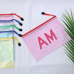 May include: A collection of colorful mesh zipper pouches. The pouches are in various colors, including pink, red, blue, green, and yellow. The pink pouch has the letters "AM" printed on it in a bold, white font. Each pouch has a colored zipper and a black zipper pull.