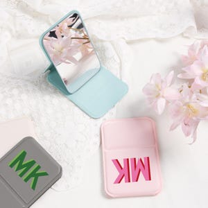 Könnte beinhalten: Ein hellblaues, zusammenklappbares Kompaktspiegel mit reflektierender Oberfläche, die rosa Blüten reflektiert. Ein rosa Kompaktspiegel mit den Buchstaben "MK" in Rot ist ebenfalls zu sehen, zusammen mit einem grauen Kompaktspiegel mit den Buchstaben "MK" in Grün.