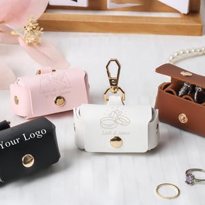 Könnte beinhalten: Vier kleine rechteckige Etuis in Schwarz, Rosa, Weiß und Braun mit goldenen Druckknöpfen. Das schwarze Etui trägt den Text Your Logo, das rosa Etui zeigt eine Strichzeichnung von Händen und das weiße Etui zeigt zwei Ringe mit dem Text Leah & James.