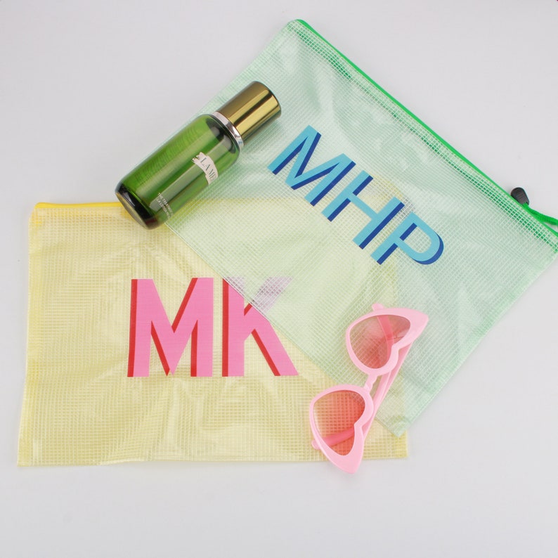May include: A clear green bottle with a gold cap, two mesh zipper pouches, and pink heart-shaped sunglasses. One pouch is yellow with the letters "MK" in pink and red. The other is light green with "MHP" in blue.