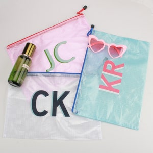 May include: Four mesh zipper pouches in pink, white, and teal, each with a different set of initials. A bottle of perfume and pink heart-shaped sunglasses are also visible. The pouches have colored zippers and are ideal for travel or organization.