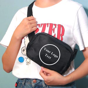 May include: A black crossbody bag with a black adjustable strap and a white circular graphic on the front containing the text Your Logo Text. The bag features a zippered closure and is worn across the chest over a white t-shirt with red lettering.