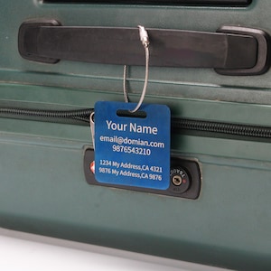 Op de afbeelding: Een blauwe bagagelabel met een zilveren metalen kabel bevestigd aan een groene koffer. Het label heeft de tekst "Your Name", "email@domian.com", "9876543210", "1234 My Address, CA 4321" en "9876 My Address, CA 9876".