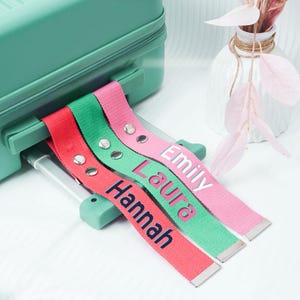 Könnte beinhalten: Drei farbige Gepäckanhänger in Rot, Grün und Rosa mit den Namen Hannah, Laura und Emily. Die Anhänger haben silberne Metallenden und Nieten. Sie sind an einem türkisfarbenen Koffer befestigt.