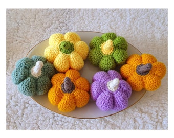 May include: Six crocheted pumpkins in various colors, including green, yellow, orange, and purple, are arranged on a white plate. The pumpkins are all about the same size and have a similar shape.