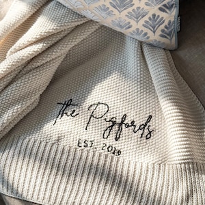 May include: Cream-colored knitted throw blanket with the embroidered text "The Pigfords EST. 2019". A decorative pillow with a blue floral pattern rests on the blanket. The blanket has a ribbed border.