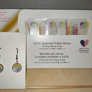 May include: A pair of silver earrings with rainbow glitter round charms. The earrings are displayed on a white card with a package of nail polish strips behind them. The nail polish strips are white with rainbow glitter and the text "100% Real Nail Polish Strips" is printed on the package.