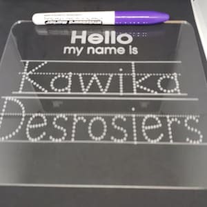May include: A clear acrylic dry-erase board with the text "Hello my name is" and the names "Kawika Desrosiers" written in dotted lines. A purple dry-erase marker is on top of the board.