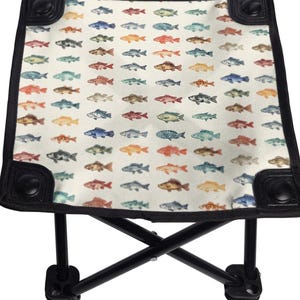 May include: A folding stool with a cream-colored seat featuring a pattern of colorful fish. The seat is bordered with black trim and has black corner accents. The black metal legs are in an X-frame design.