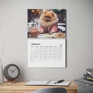 Peut inclure: Un calendrier mural pour f&eacute;vrier 2027 pr&eacute;sentant une photographie d'un chien Pom&eacute;ranien au pelage duveteux portant une veste rose &agrave; col en polaire blanc, assis &agrave; une table avec une tasse rose et une assiette de p&acirc;tes dans un d&eacute;cor enneig&eacute;.