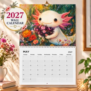 Peut inclure: Un calendrier mural 2027 illustré d'un axolotl blanc tenant un bouquet de fleurs. La partie supérieure montre l'axolotl dans un jardin coloré, tandis que la partie inférieure présente la grille du calendrier de mai 2027 avec les jours.