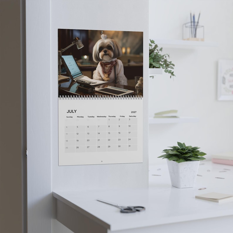 Peut inclure: Un calendrier mural pour juillet 2027 pr&eacute;sentant une photographie d'un petit chien &agrave; poils longs portant une &eacute;charpe &agrave; motifs, assis &agrave; un bureau avec un ordinateur portable et une lampe. Le calendrier a une reliure spirale blanche en haut.