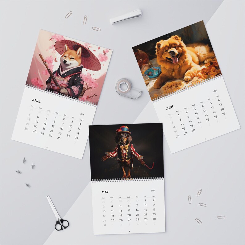 Peut inclure: Un calendrier mural 2026 avec trois pages ouvertes affichant les mois d'avril, mai et juin. Chaque page pr&eacute;sente une illustration de chien diff&eacute;rente, dont un Shiba Inu en kimono, un teckel en costume de ma&icirc;tre de cirque et un chow-chow.