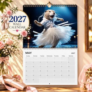 Peut inclure: Un calendrier mural 2027 représentant un chien de prairie portant un tutu blanc et des chaussons de danse. La page du calendrier affiche le mois de mai 2027 avec une grille de dates. Le texte 2027 WALL CALENDAR est orné de roses roses.