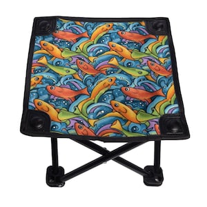 May include: A folding stool with a colorful fish pattern. The seat features orange, yellow, and green fish swimming in blue and purple waves. The frame is black, and the stool is designed for portability and outdoor use.