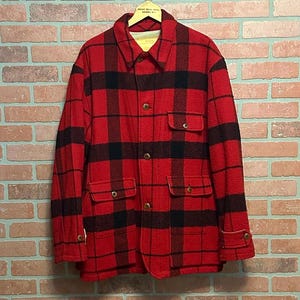May include: A red and black plaid jacket with four front pockets and button closures. The jacket is hanging on a wooden hanger against a brick wall background. The jacket has a collared neckline.