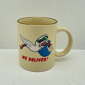 Può includere: Tazza in ceramica color crema con bordo e manico marroni. La tazza presenta una cicogna dei cartoni animati con una maschera verde e che trasporta un bambino in un fagotto blu. Il testo "WE DELIVER!" è stampato in rosso.
