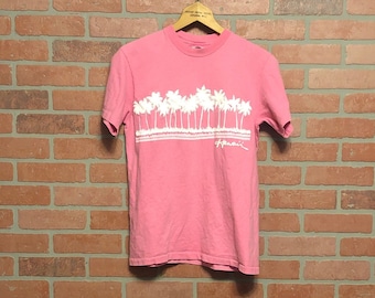 May include: Pink t-shirt featuring a white palm tree design and the word "Hawaii". The shirt is hanging on a wooden hanger against a brick wall.
