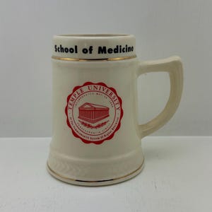 May include: Cream-colored ceramic mug with a handle, featuring the text "School of Medicine" in black and gold trim. The mug displays a red and white Temple University emblem. A classic stein-style design.
